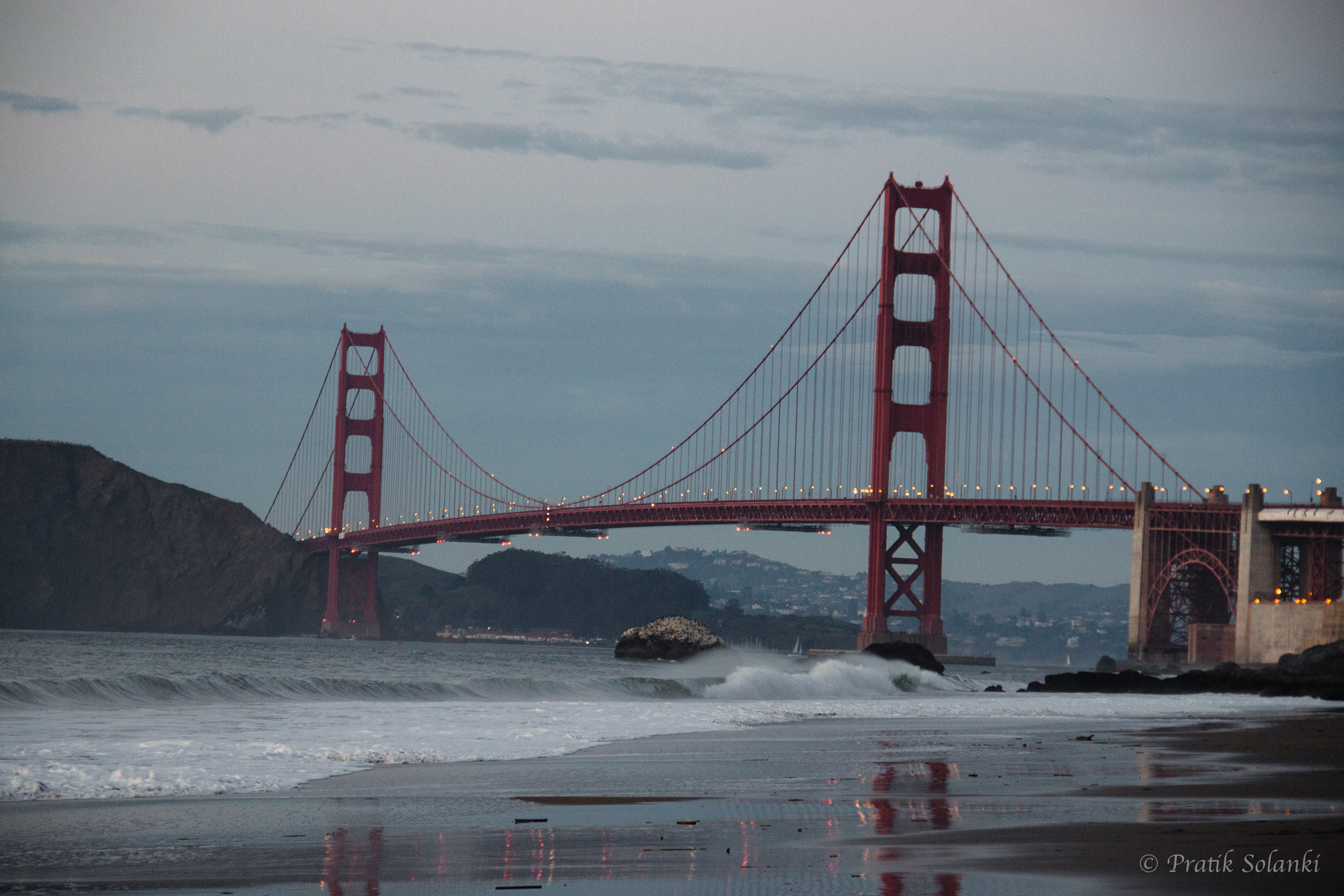 Golden Gate Bridge lit at dusk, viewed from Baker Beach with waves in the foreground